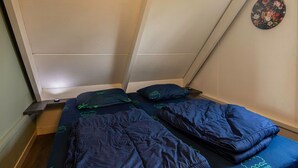 Une petite pièce aux murs inclinés, contenant un lit, un oreiller et un objet rond fixé au mur.