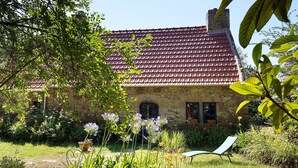 A stone building with a red-tiled roof, a garden with various plants, and a chair in the foreground.
