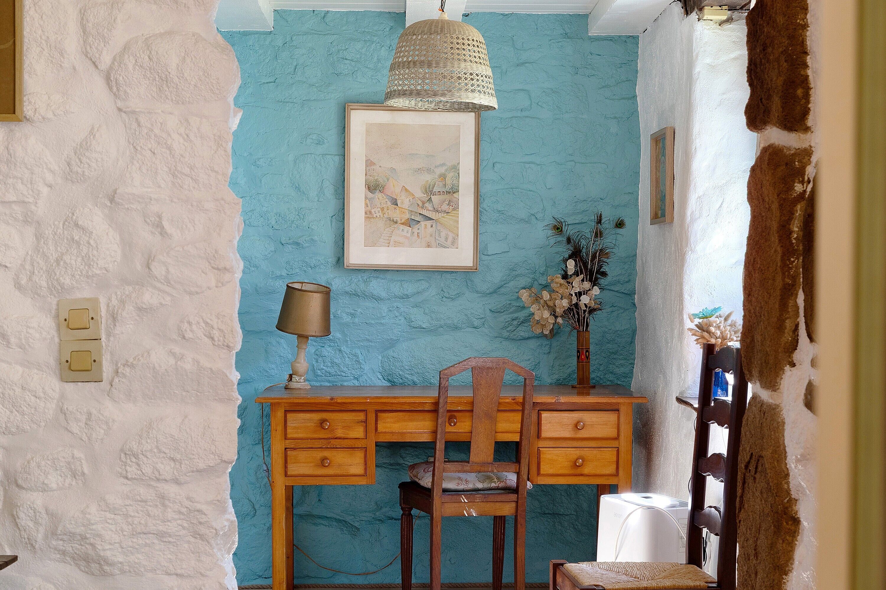 A room with a blue textured wall, a wooden desk with drawers, a chair, a hanging lamp, and a framed picture.