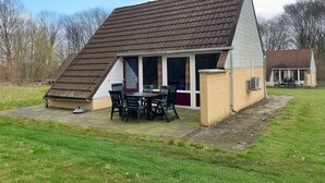Ein kleines, eingeschossiges Gebäude mit einem geneigten Dach, einer Terrasse mit Stühlen und einem Fenster mit Blick auf eine Wiese.