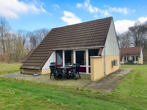 A small, single-story building with a sloped roof, a patio with chairs, and a window overlooking a grassy area. - Bungalowpark Simpelveld - Alpha 75 by Interhome (Simpelveld)