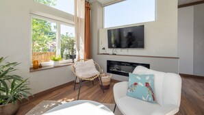 A modern living room with a fireplace, a TV mounted on the wall, a white sofa, a wicker chair, and a large window with curtains.