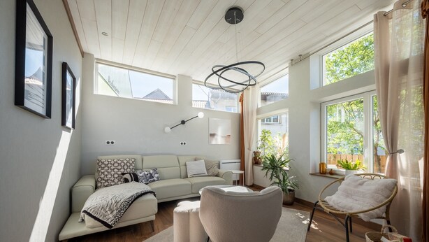 A modern living room with a sofa, armchair, and ottoman, large windows, and a ceiling light fixture.