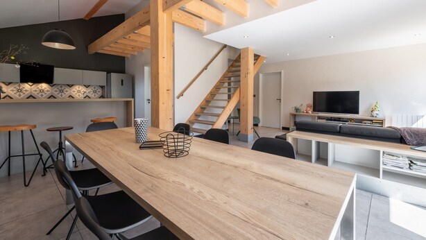 A modern kitchen with wooden beams, a long dining table, and bar stools. - Grand Gîte Les Hangars: South-Facing Terrace, 4 Bedrooms, Sauna & Whirlpool – Beds Made, Cleaning Included (Saulxures-sur-Moselotte)