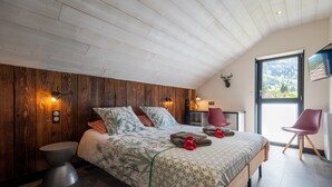 A bedroom with a wooden headboard, a bed with patterned bedding, a round side table, a chair, a television, and a window with a view. - Grand Gîte Les Hangars: South-Facing Terrace, 4 Bedrooms, Sauna & Whirlpool – Beds Made, Cleaning Included (Saulxures-sur-Moselotte)