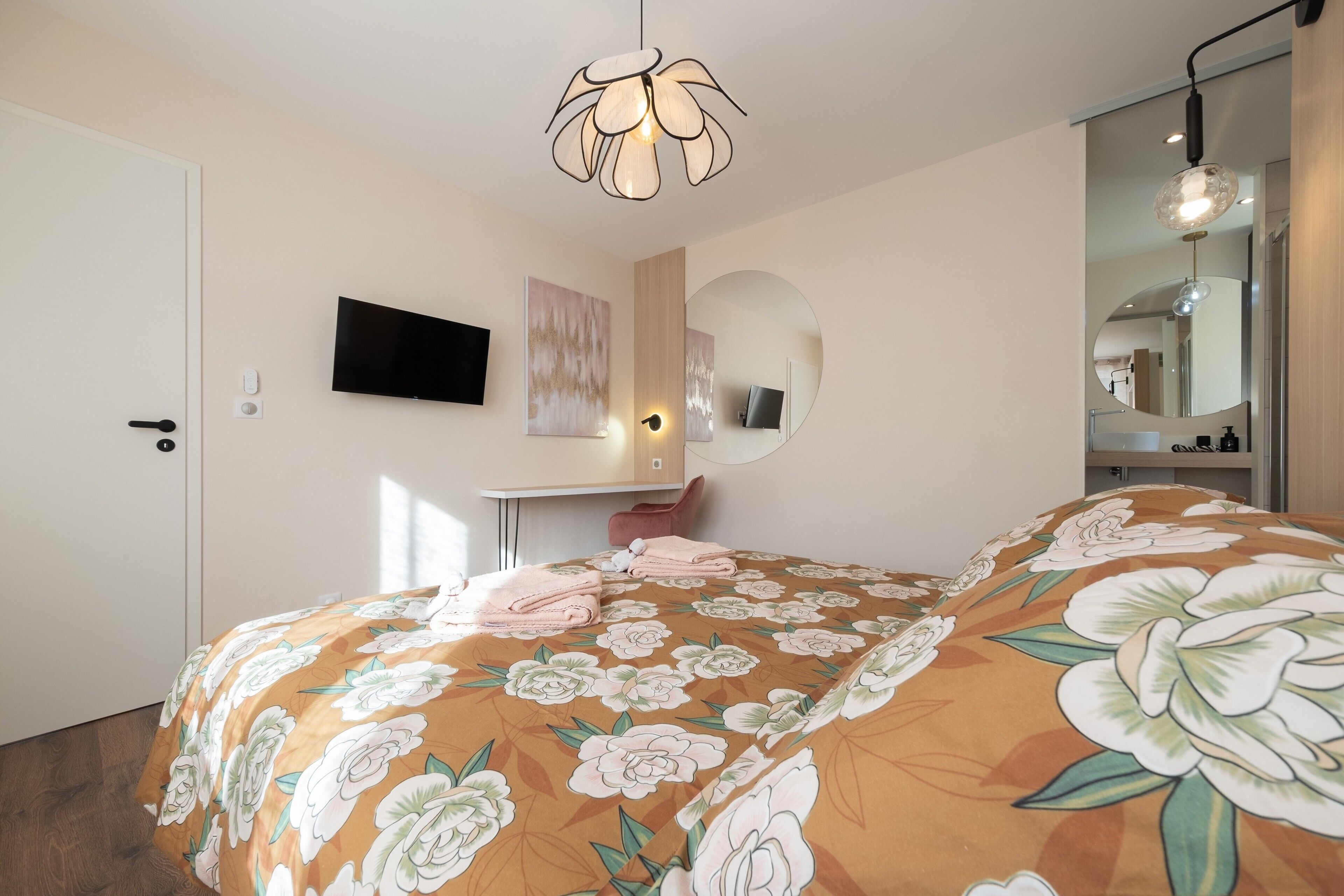 Une chambre à coucher avec une couvre-lit à motifs floraux, un téléviseur fixé au mur, une table de chevet, un miroir et une porte.