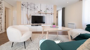 Un soggiorno moderno con una televisione a schermo piatto, una poltrona bianca, un divano color petrolio, un tavolino in legno e una parete decorata con motivi di uccelli.