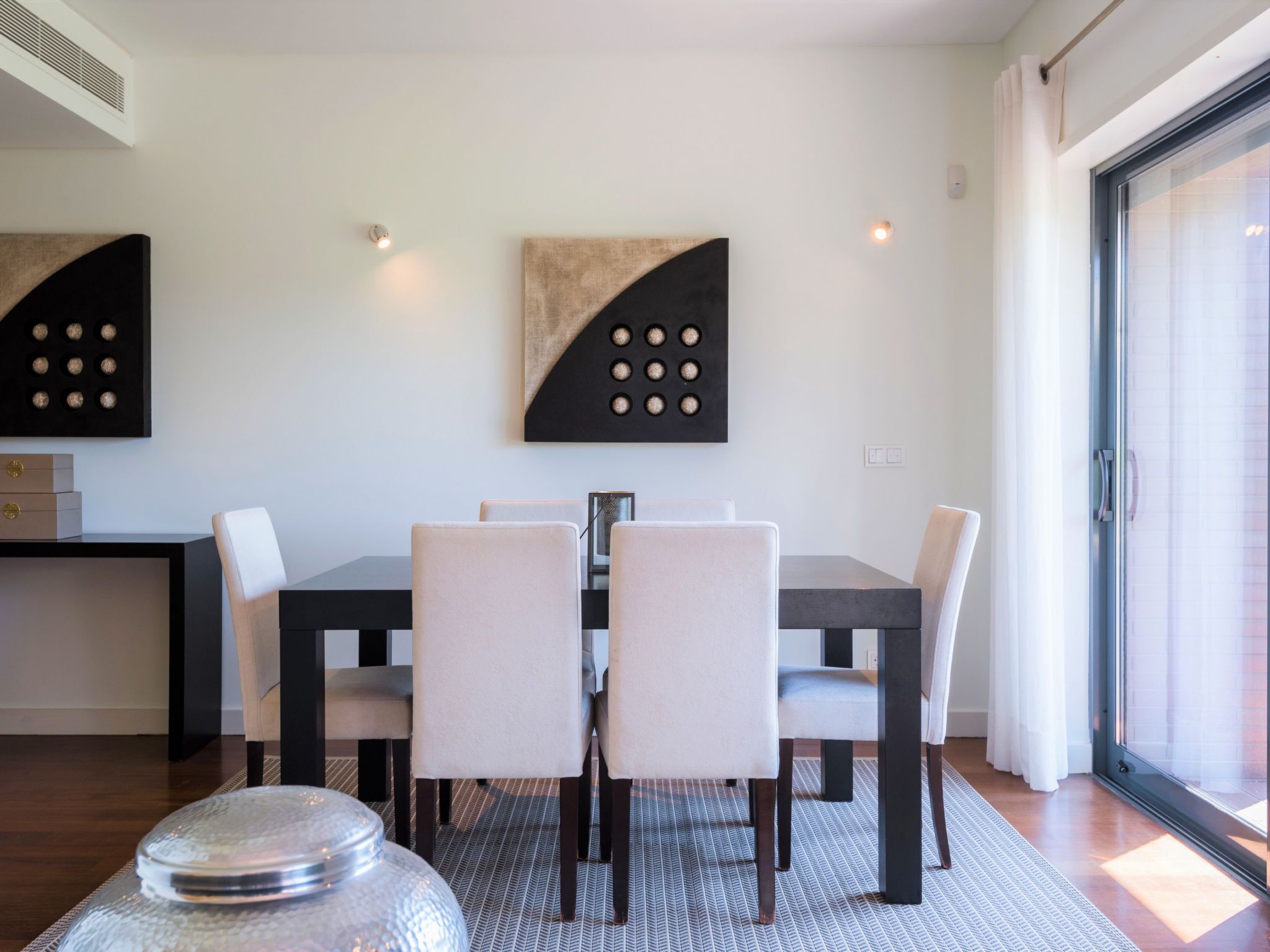 A dining area with a table and chairs, a decorative wall piece, and a large window.