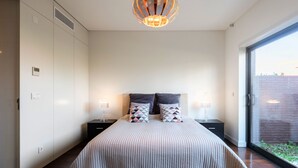 Ein Schlafzimmer mit einem großen Bett, zwei Nachttischen, einer modernen Pendelleuchte und einer Schiebeglastür, die zu einem Außenbereich führt.