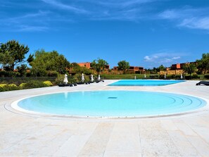 A large outdoor swimming pool with lounge chairs and umbrellas, surrounded by greenery and buildings. - Apartment  by Interhome (Quarteira)