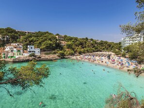 Una playa costera con aguas turquesas cristalinas, una orilla arenosa y edificios al fondo.