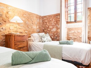 A room with a stone wall, a wooden nightstand, a lamp, two beds with white bedding, and a window with curtains. - Casa Antigua by Interhome (Santanyí)