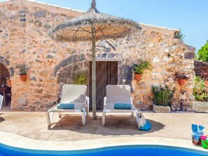 A poolside area with two lounge chairs, a thatched umbrella, and a stone building with potted plants. - Casa Antigua by Interhome (Santanyí)