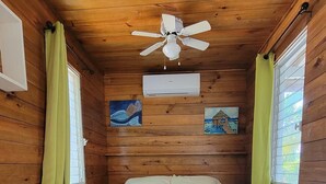 Une pièce de cabane en bois comprenant un lit, un ventilateur de plafond et deux fenêtres avec des rideaux.
