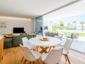 Ein modernes Wohnzimmer mit großem Fenster, einem Flachbildfernseher, einem grünen Sofa und einem Essbereich mit weißen Stühlen und einem runden Tisch.