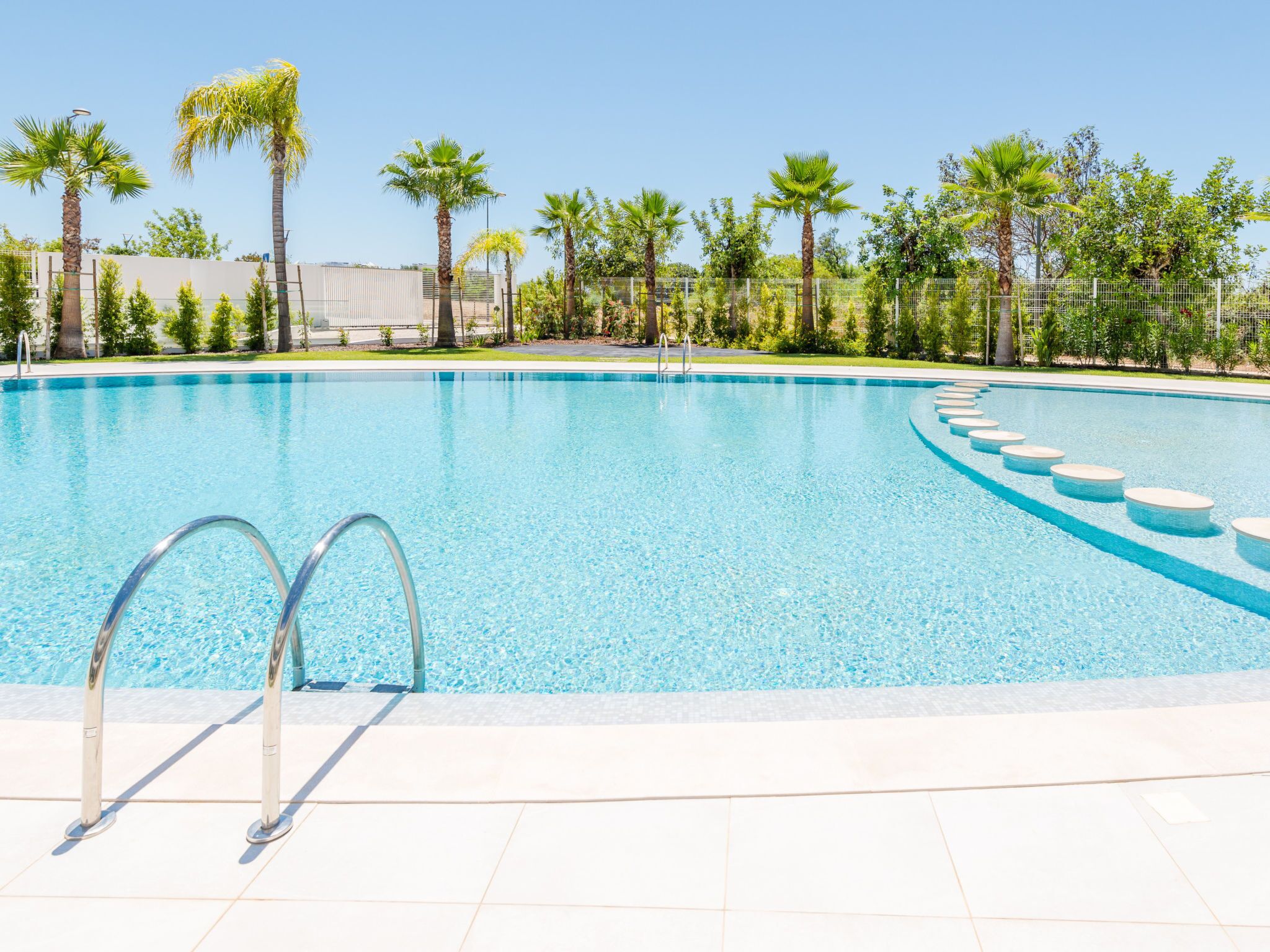 Une piscine d’un bleu éclatant, avec des marches menant à l’intérieur, entourée de palmiers et d’une clôture.