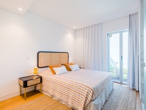 Ein modernes Schlafzimmer mit einem großen Bett, einem Nachttisch mit Lampe und Blick ins Freie durch eine Schiebetür.