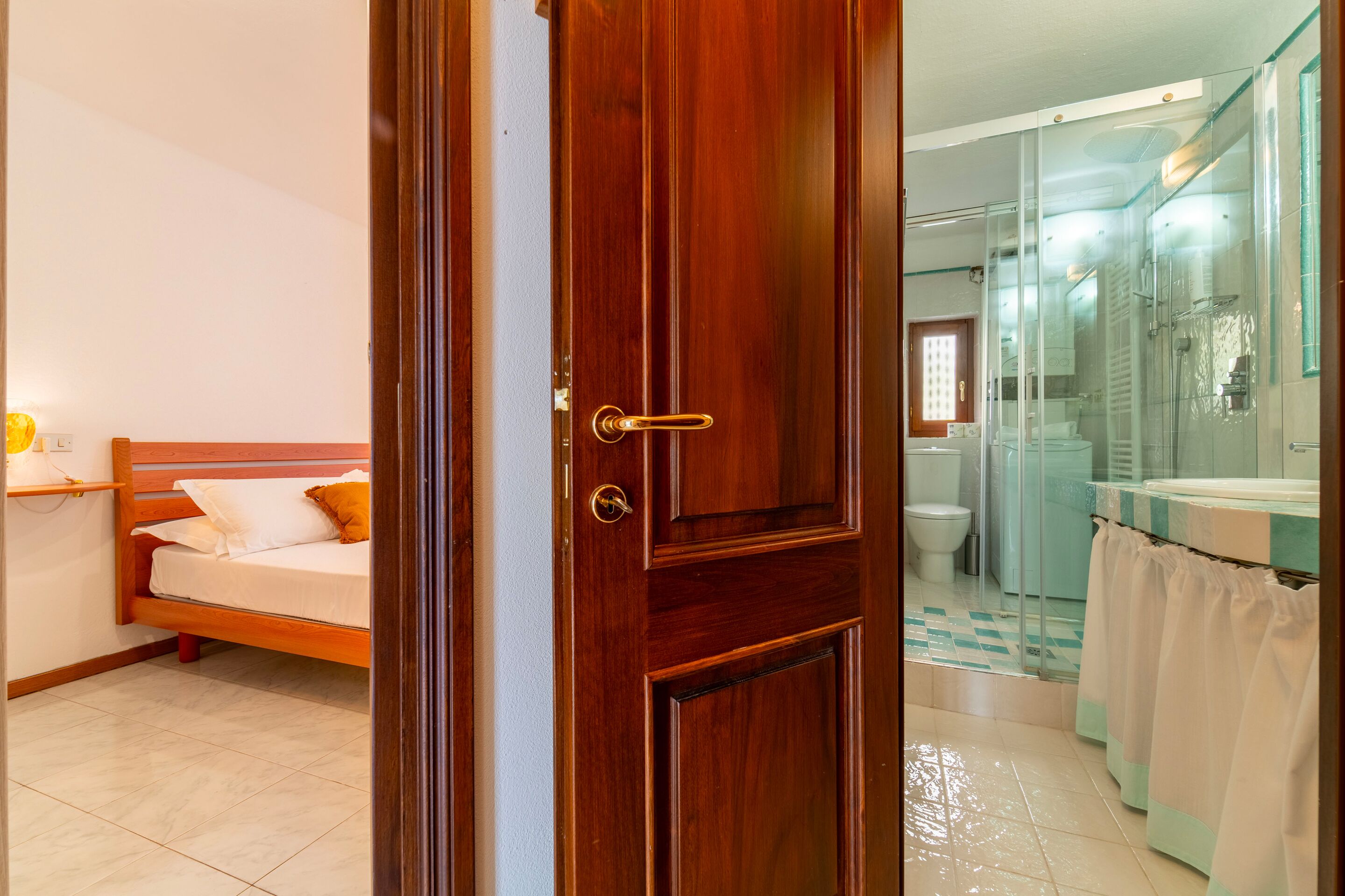 A hotel room with a wooden door, a bed, a bathroom with a glass shower enclosure, and a visible toilet.