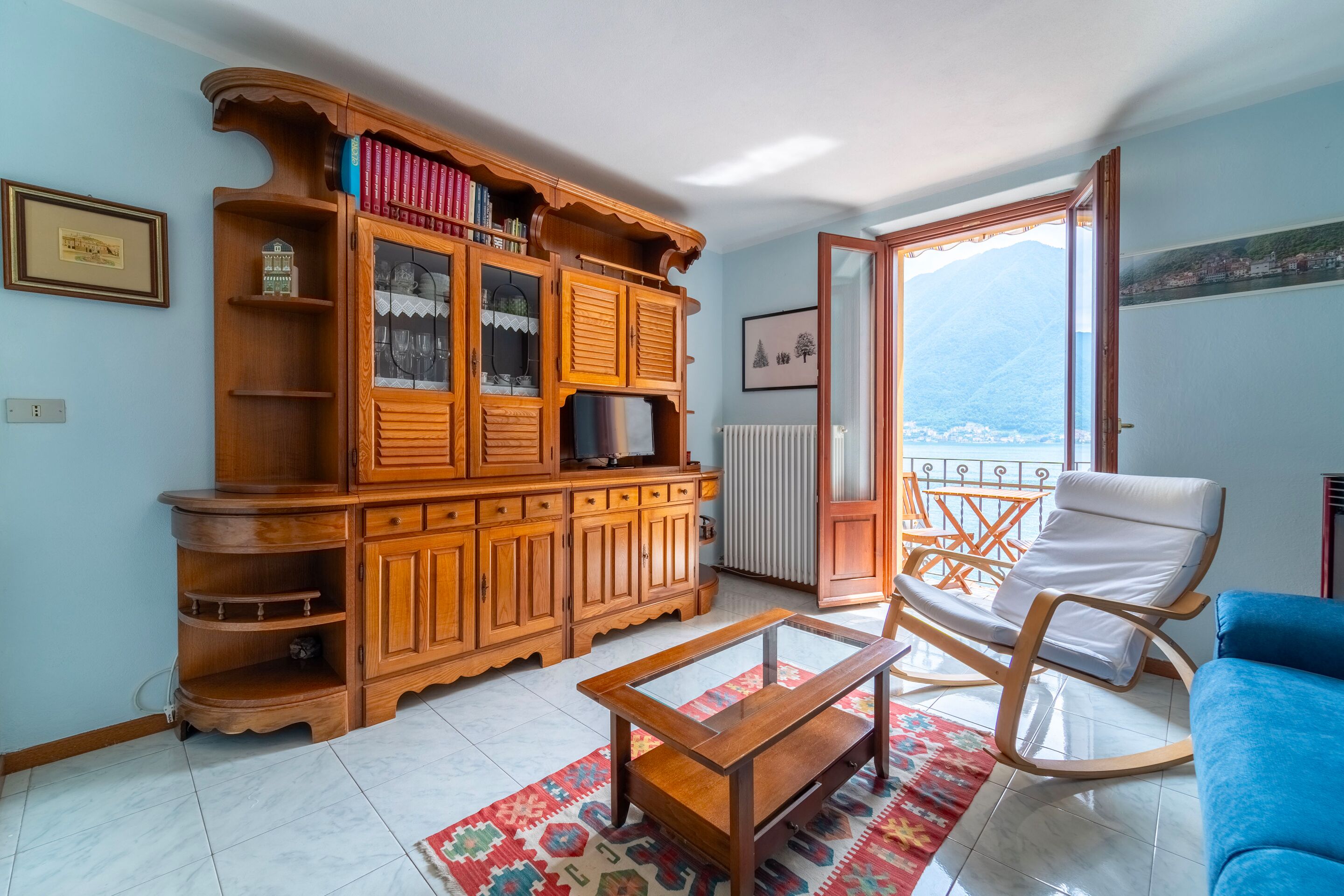 Ein Wohnzimmer mit Holzschrank, Glastisch, blauem Sofa, Schaukelstuhl und Bergblick durch eine Balkontür.