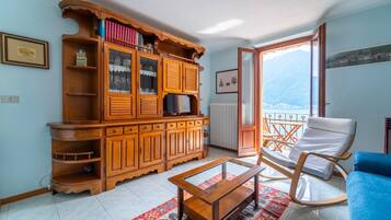 Un salon avec un meuble en bois, une table basse en verre, un canapé bleu, un fauteuil à bascule et une vue sur les montagnes, visible à travers une porte-fenêtre donnant sur un balcon.