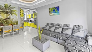 A modern living room with a sectional sofa, a dining area with chairs and a table, a kitchen area with yellow cabinets, and a tropical-themed wall mural. - Jaddoo Apartment at Azeer Residence (Grand-Baie)