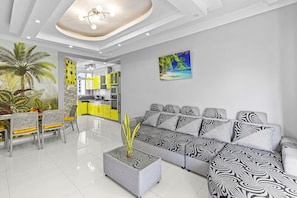 A modern living room with a sectional sofa, a dining area with chairs and a table, a kitchen area with yellow cabinets, and a tropical-themed wall mural. - Jaddoo Apartment at Azeer Residence (Grand-Baie)