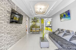 A modern living room with a brick wall, a flat-screen TV, a patterned sofa, a dining area with a table and chairs, and a kitchen with yellow cabinets. - Jaddoo Apartment at Azeer Residence (Grand-Baie)