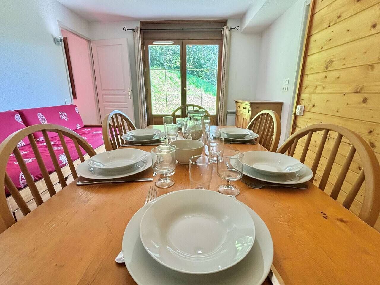Une table à manger dressée pour six personnes, avec des chaises, une fenêtre avec des rideaux et une porte sur le côté gauche.