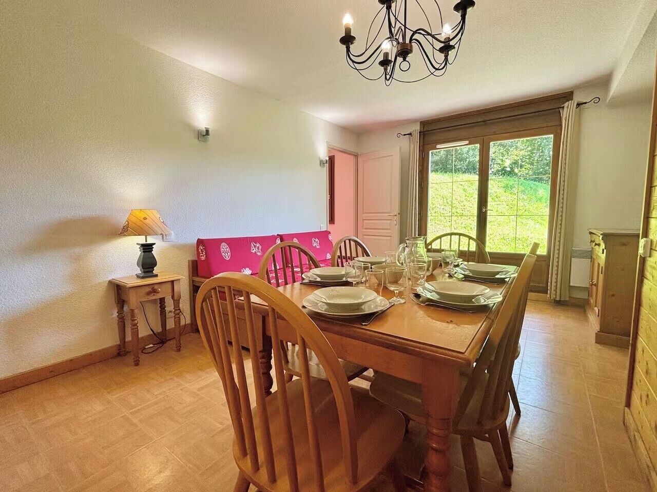A dining area with a wooden table set for four, chairs, a chandelier, and a view through a door to a garden.