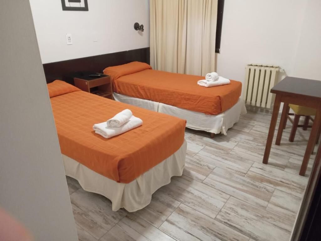 A hotel room with two beds, a wooden table, a nightstand, a radiator, and a window with curtains.