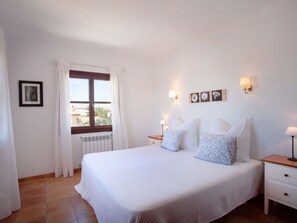 A hotel room with a large bed, two bedside tables, a window with curtains, and wall-mounted lamps. - Sa Bassa by Interhome (Cala Llombards)