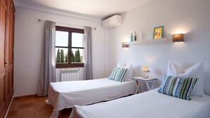 Ein Hotelzimmer mit zwei Betten, einem Fenster mit Vorhängen, einem Heizkörper, einer Wand-Klimaanlage und einem kleinen Tisch mit einer Lampe.