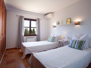 Ein Hotelzimmer mit zwei Betten, einem Fenster mit Vorhängen, einem Heizkörper, einer Wand-Klimaanlage und einem kleinen Tisch mit einer Lampe.