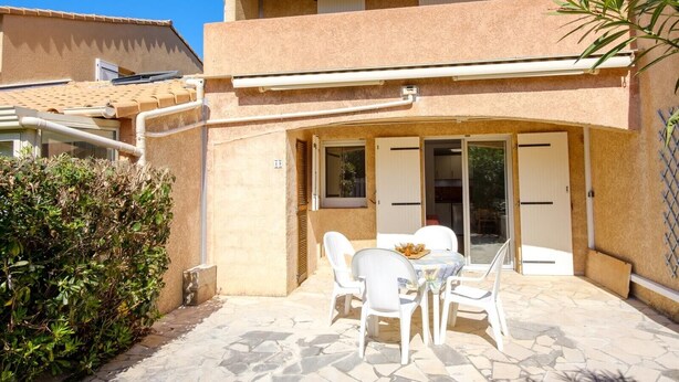 Ein zweistöckiges Gebäude mit einer Terrasse, die einen Tisch und Stühle sowie einen kleinen Gartenbereich mit Büschen umfasst. Ein Glastürschieber dient als Eingang.