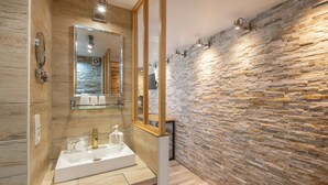 A modern bathroom with a stone wall, a large mirror, and a white sink with gold fixtures. - Studio near Old Port, Panier and Station (Marseille)