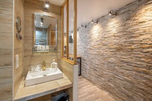 Ein modernes Badezimmer mit Steinwand, großem Spiegel und weißem Waschbecken mit goldfarbenen Armaturen.
