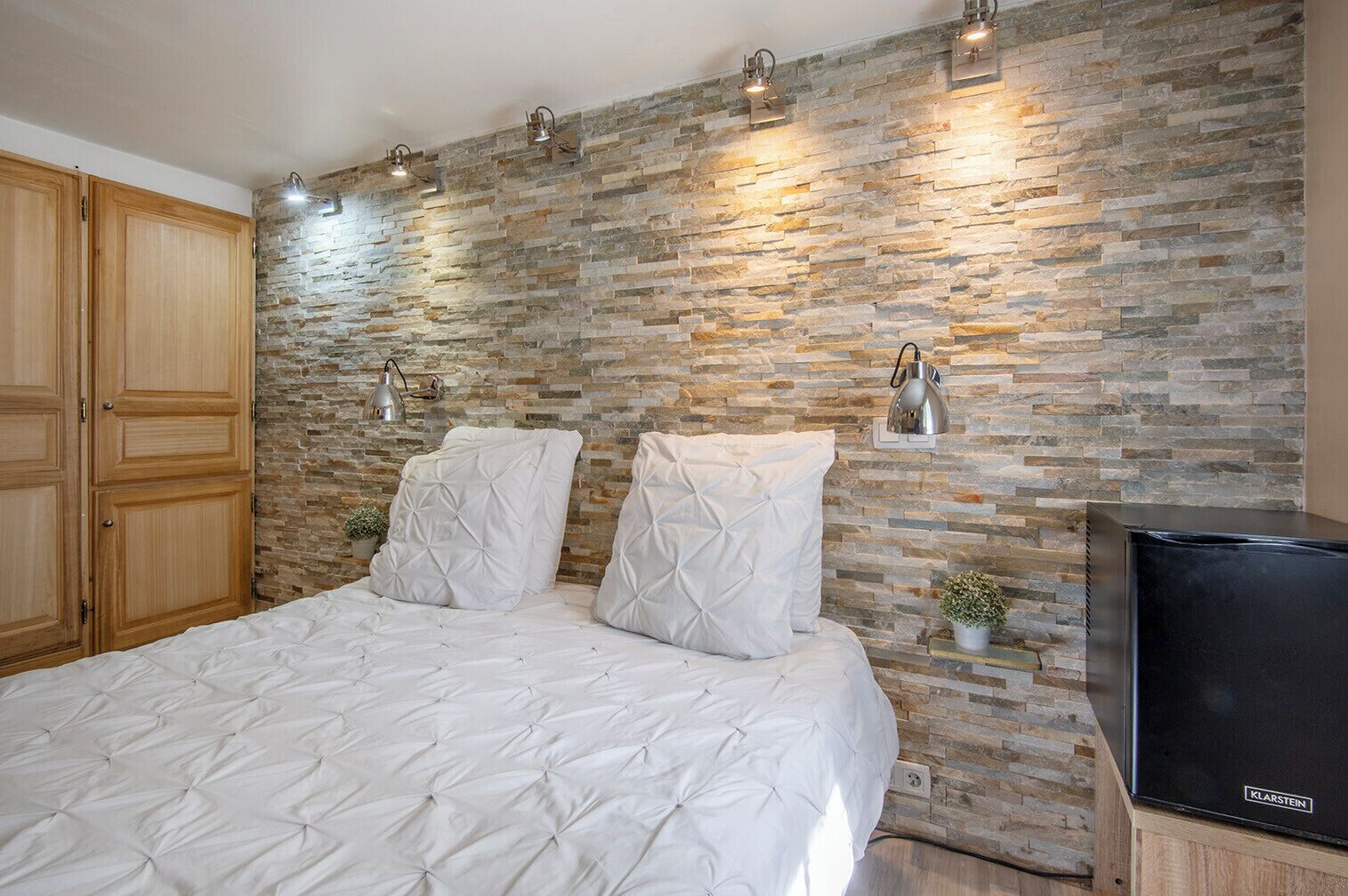 Une chambre avec un mur en pierre, un lit avec une literie blanche, une armoire en bois et un téléviseur posé sur un meuble en bois.