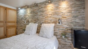 A bedroom with a stone wall, a bed with white bedding, wooden wardrobe, and a TV on a wooden stand. - Studio near Old Port, Panier and Station (Marseille)
