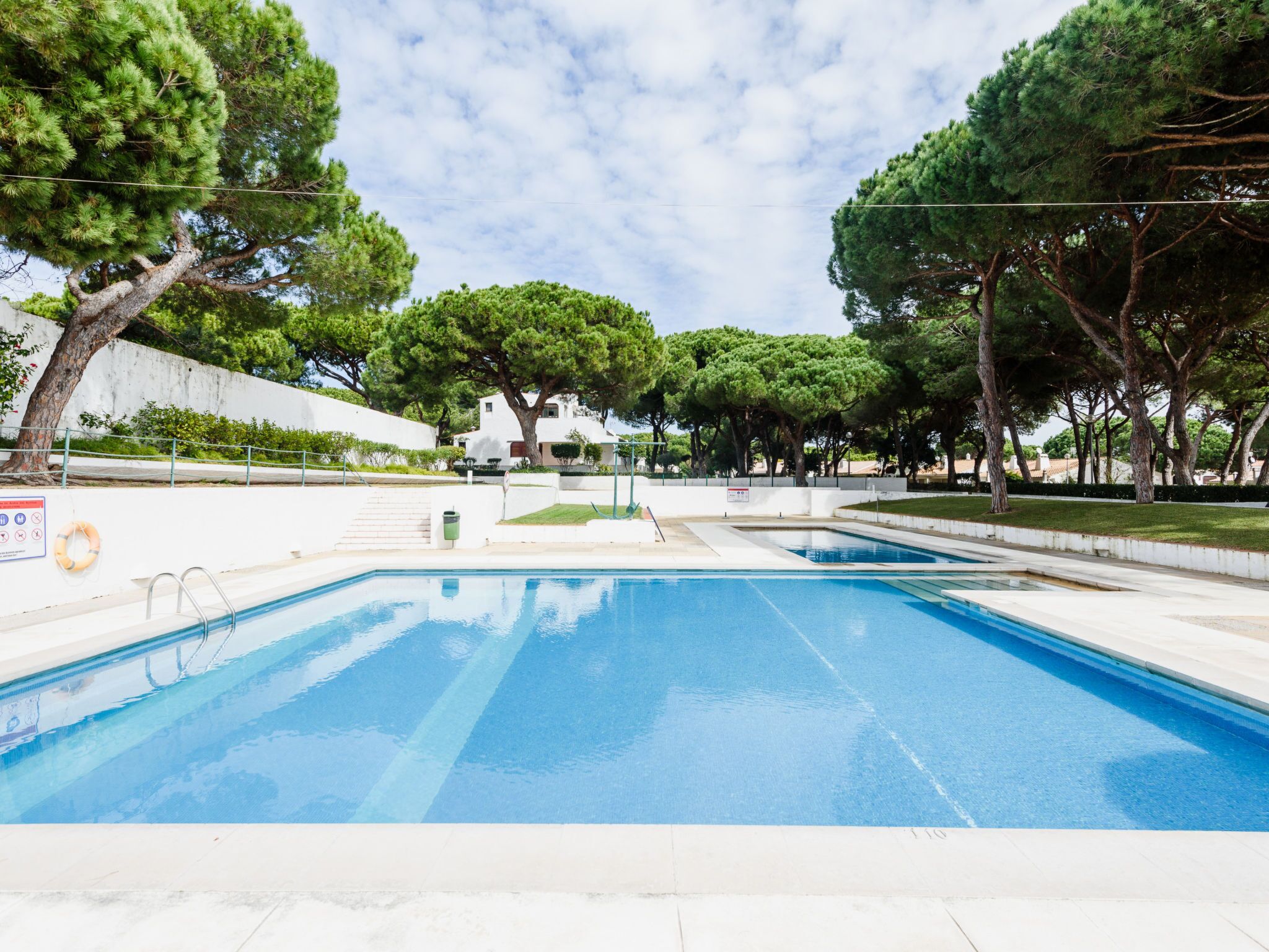 Una piscina con agua azul cristalina, rodeada de árboles y un edificio blanco al fondo.