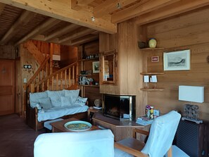 Ein gemütliches Blockhaus-Interieur mit einer Holztreppe, einer Couch, einem Esstisch mit Teller, einem Fernseher und einem Kamin.