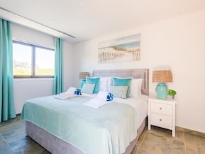 A neatly made bed with a light blue bedspread and pillows, a bedside table with a lamp and a plant, and a window with curtains. - Apartment  by Interhome (Albufeira)