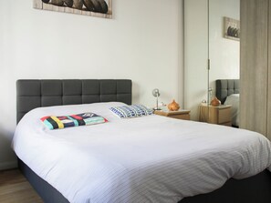 Ein ordentlich bezogenes Bett mit grauem Polsterkopfteil, ein Nachttisch mit Lampe und eine kleine Vase.