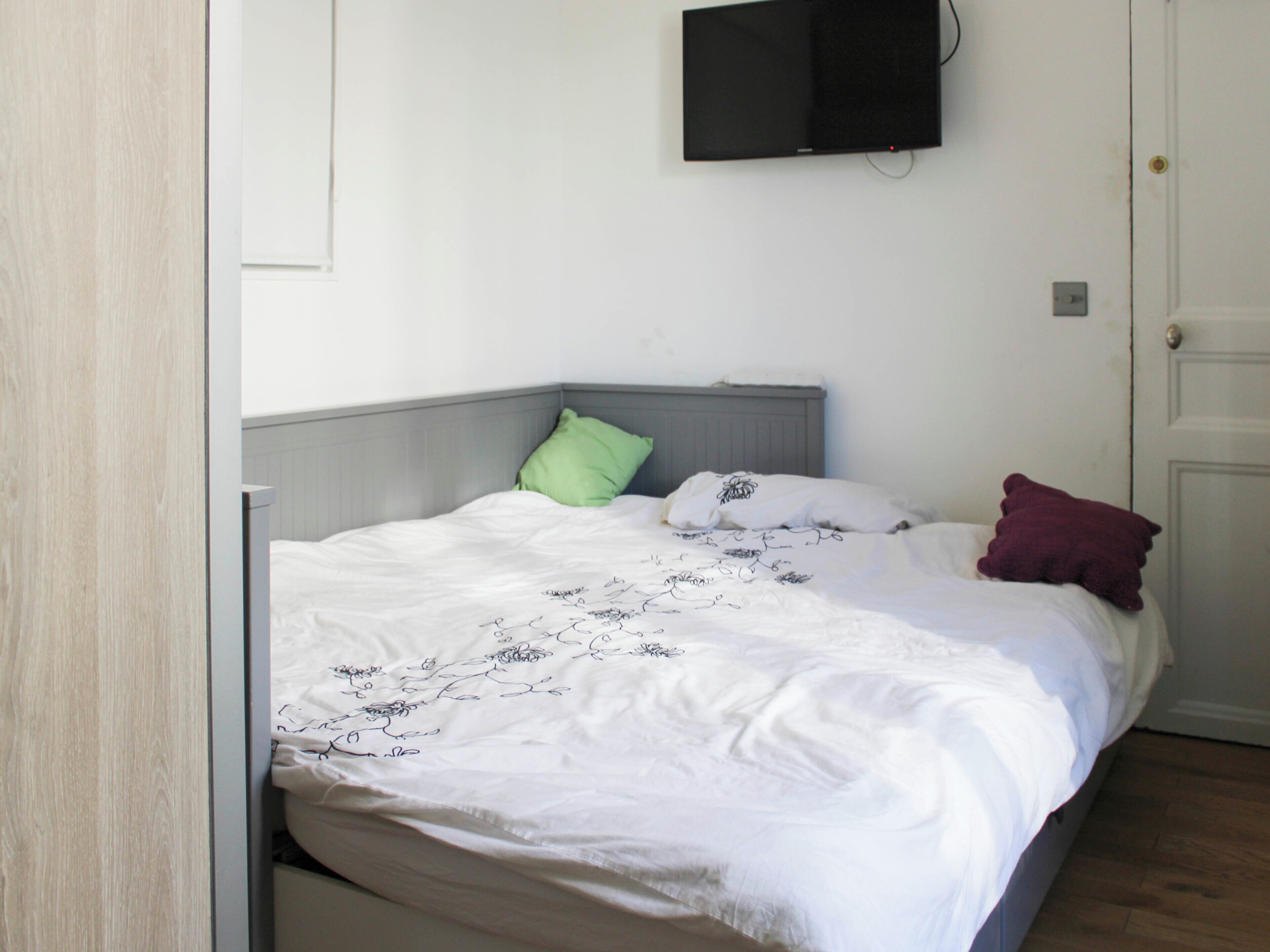 A bed with a white bedspread and pillows, a grey headboard, a mounted television, and a door.