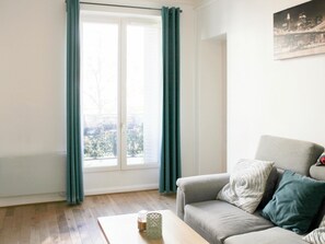 A living room with a sofa, a coffee table, and a window with curtains. - 2-bedroom apartment - Eiffel Tower (Paris)
