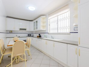 A kitchen with white cabinets, a dining table with chairs, a microwave, and a window with decorative bars. - Apartment  by Interhome (Vilamoura)