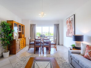 A modern living room with a dining area, a sofa, a lamp, and a large window with curtains. - Apartment  by Interhome (Vilamoura)