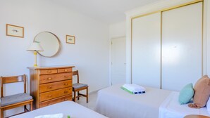 Quarto de hotel com duas camas, uma cômoda de madeira, uma cadeira, um espelho e um guarda-roupa.