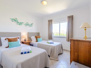 A hotel room with two beds, a nightstand, a lamp, a window with curtains, and a view of buildings outside. - Apartment  by Interhome (Vilamoura)