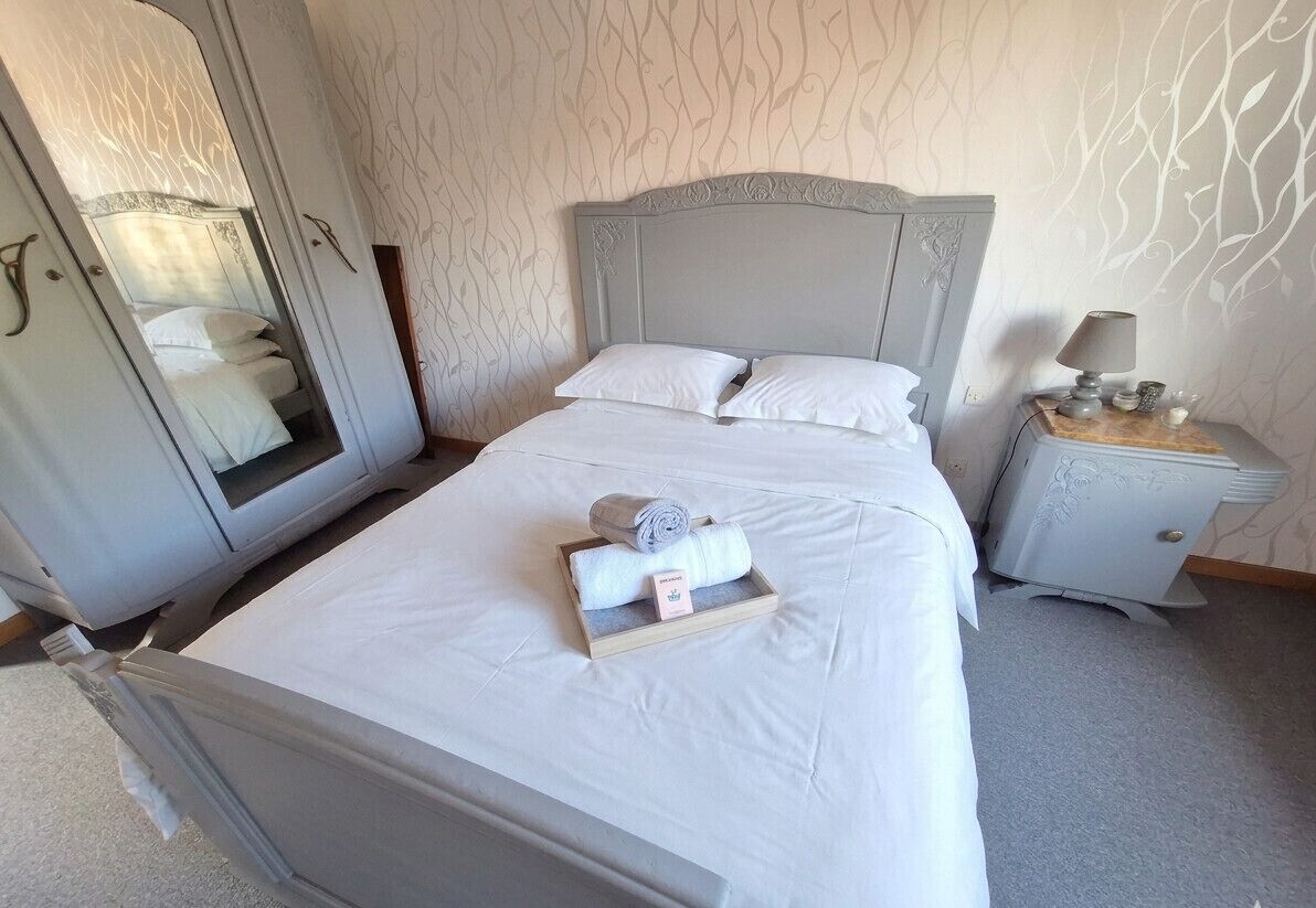 Een net opgemaakt bed met witte lakens, een houten nachtkastje met een lamp en een spiegel aan de muur.