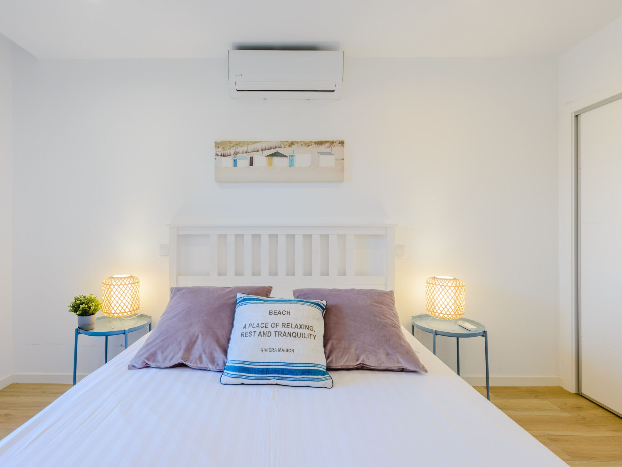 A bedroom with a white bed, two nightstands with lamps, a painting above the bed, and an air conditioning unit on the wall.